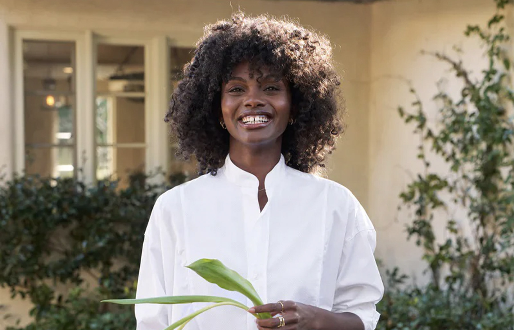 A woman with curly hair, wearing a white blouse, smiles while holding a green plant. She stands in front of a house with a pale facade and leafy greenery around the doorway. Fearrington Village