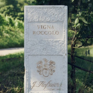 A stone marker in a vineyard reads "Vigna Roccolo" with the name "J. Hofstätter" beneath. It is surrounded by lush green vines and grass. Fearrington Village