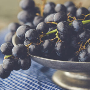 A close-up of a silver bowl filled with dark purple grapes, resting on a blue and white checkered cloth. The grapes have a fresh and glossy appearance, and light softly highlights their texture. Fearrington Village