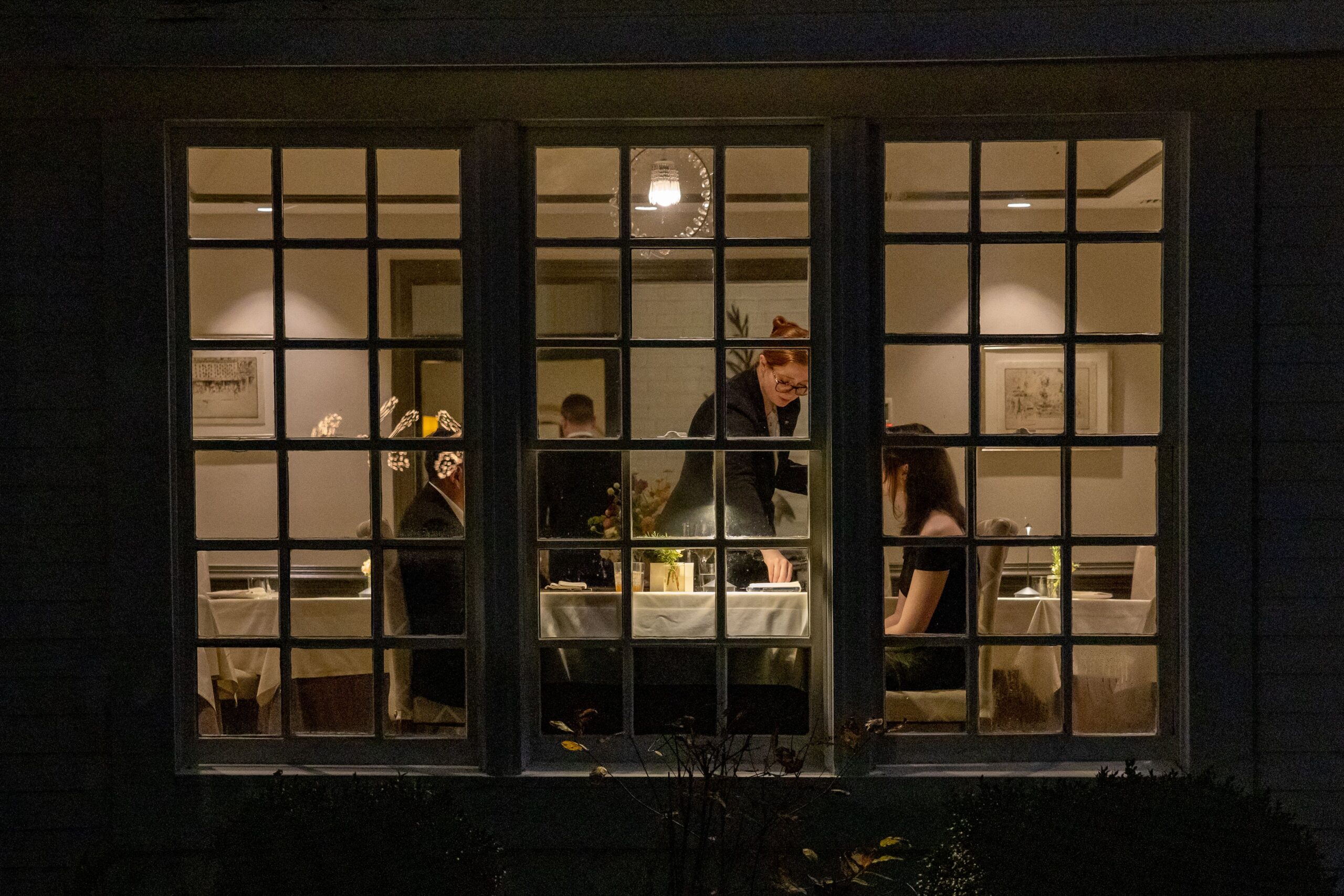 A warmly lit restaurant interior viewed through a window, where a waiter attends to a seated couple at a table. The setting is cozy, with decorative wall art and ambient lighting enhancing the intimate atmosphere. Fearrington Village