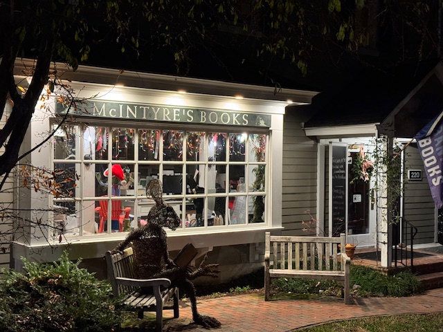 A cozy bookstore named "McIntyre's Books" is lit up at night. The window displays various books and decorations. A whimsical sculpture of a seated figure is on the sidewalk in front, next to wooden benches. A flag outside reads "Books. Fearrington Village