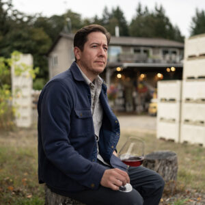 A man in a blue jacket sits outdoors on a tree stump, holding a glass of red wine. Behind him is a house surrounded by trees and stacked crates. The setting conveys a rustic, peaceful atmosphere. Fearrington Village