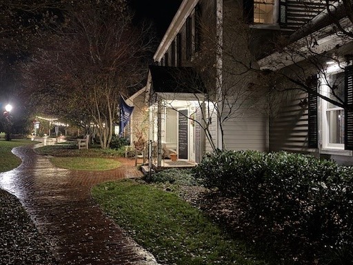 A nighttime scene shows a cozy, well-lit house with a front porch. The house is surrounded by trees and shrubs, and a curved brick pathway glistens with moisture, leading visitors through the landscaped yard. Fearrington Village