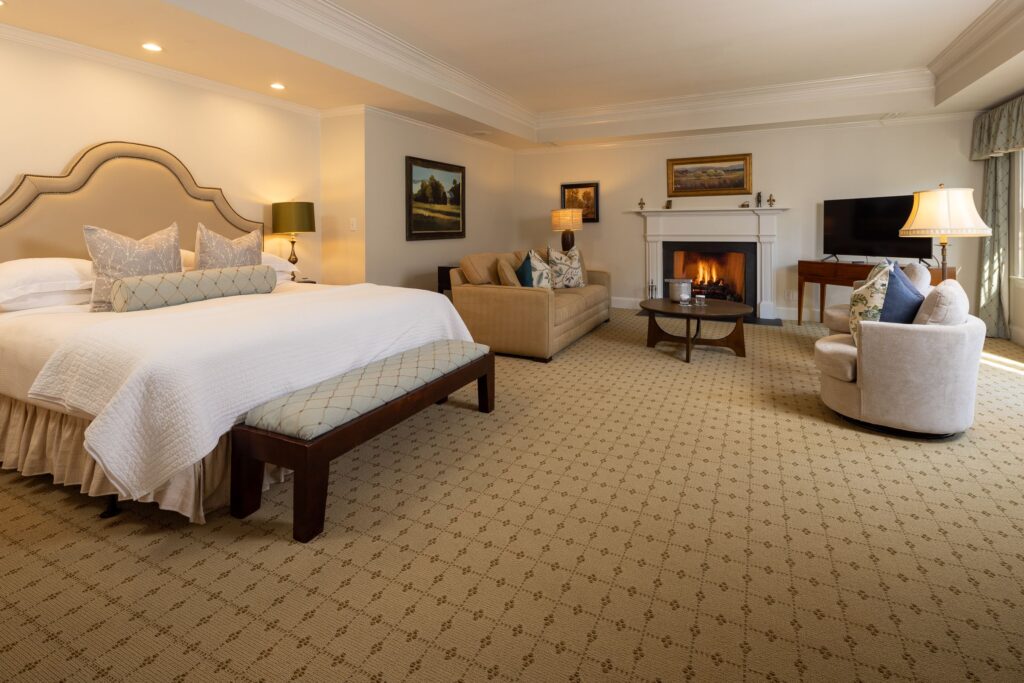 A spacious hotel room with a king-sized bed, a sofa, armchairs, a fireplace, and a flat-screen TV. The room is warmly lit, featuring elegant decor, a beige carpet, and framed artwork on the walls. A coffee table is placed near the seating area. Fearrington Village