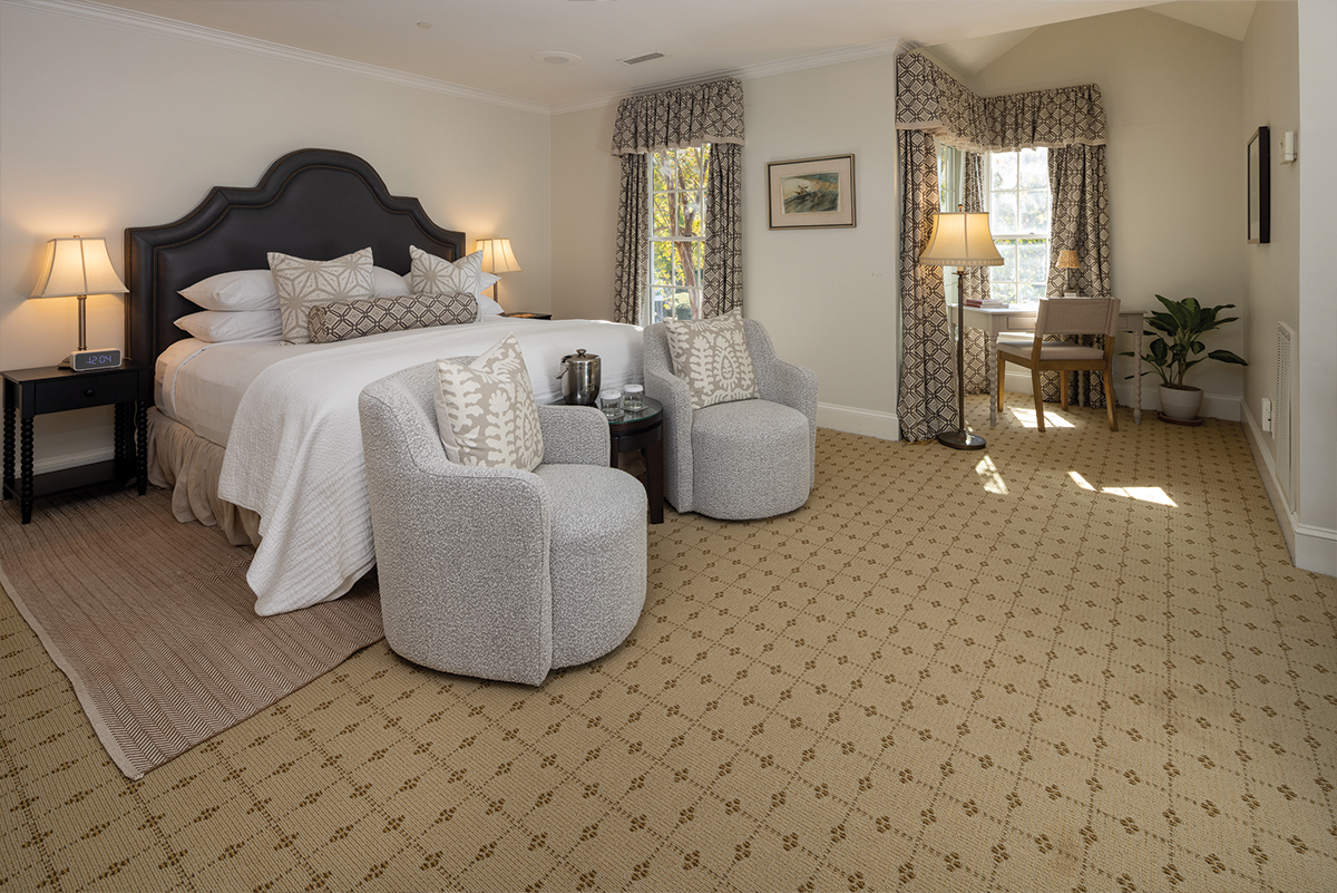 A cozy hotel room with a large bed adorned with cushions and pillows. Two soft gray chairs face the bed next to a small table with a drink set. Natural light streams in from a window, illuminating a desk by the corner and lamps on each side. Fearrington Village