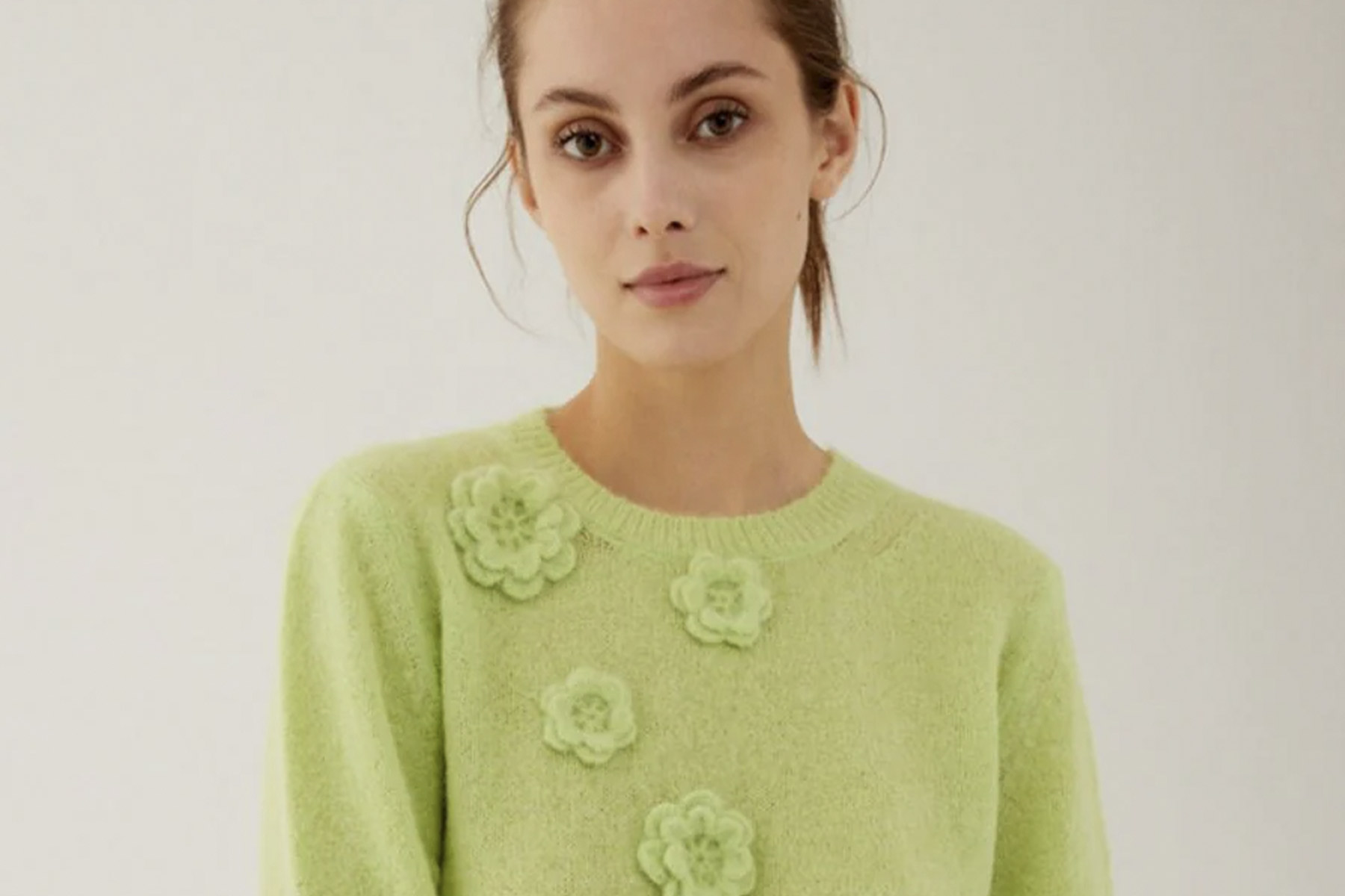 A person wearing a light green sweater with flower embellishments on the front stands against a neutral background. Fearrington Village