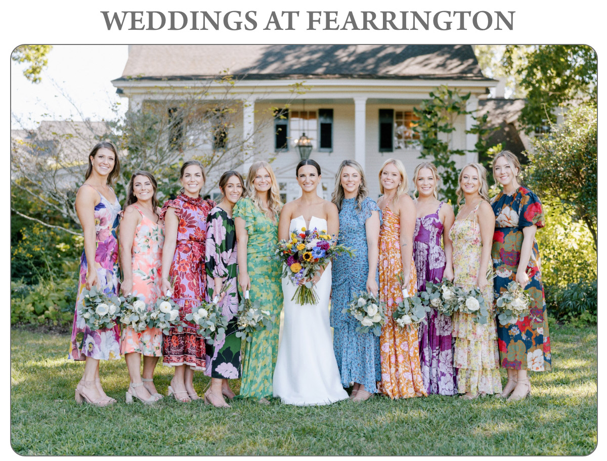 A bride in a white dress stands with 11 bridesmaids wearing vibrant floral dresses, holding bouquets. They pose on a grassy lawn in front of a house. Text above reads "Weddings at Fearrington. Fearrington Village