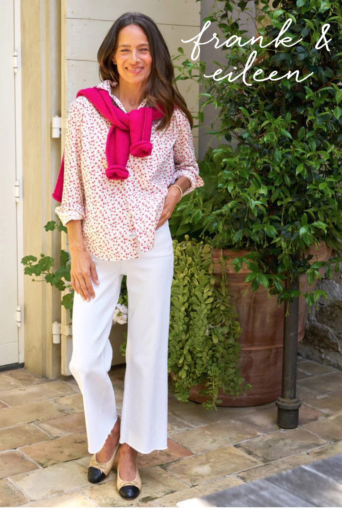 A woman stands outdoors on a stone patio. She is wearing a pink-patterned shirt, white pants, and has a pink sweater draped over her shoulders. Her hair is long and brown. There are green plants behind her, and "frank & eileen" is written in the top right corner. Fearrington Village