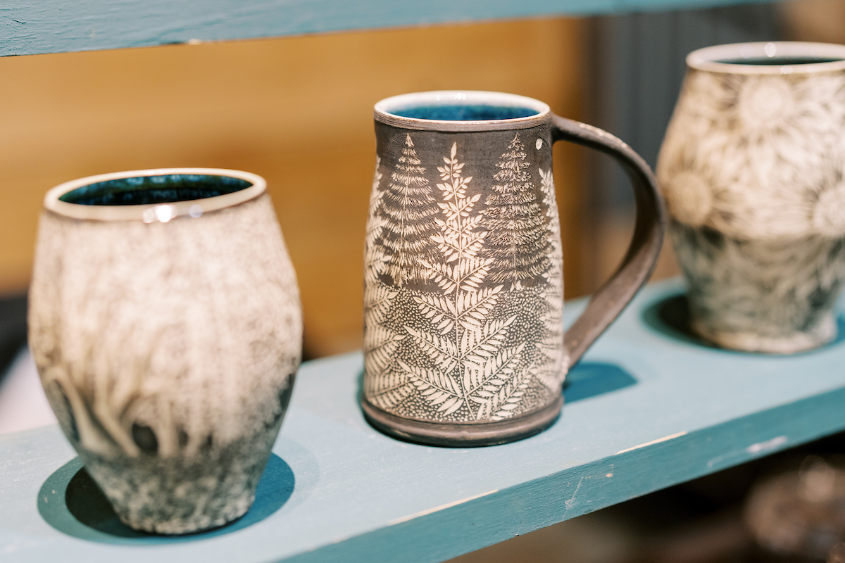 Three ceramic mugs with detailed etched designs, featuring natural patterns like trees and leaves, displayed on a shelf. The central mug is taller with a handle, and the two on either side lack handles. The colors are earthy and muted. Fearrington Village