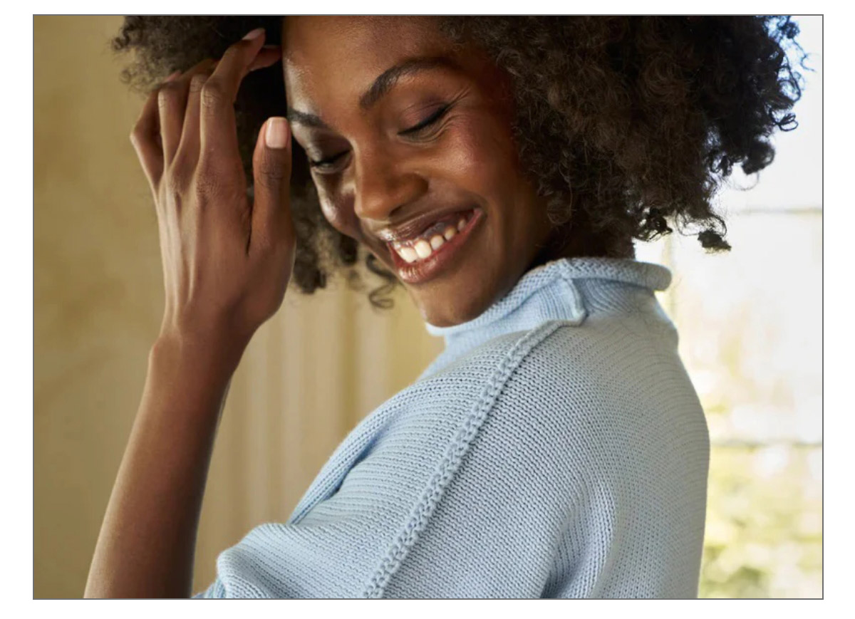 A person with curly hair smiles while glancing down. They are wearing a light blue knit sweater and are indoors in a softly lit room. One hand is raised near their face. Fearrington Village