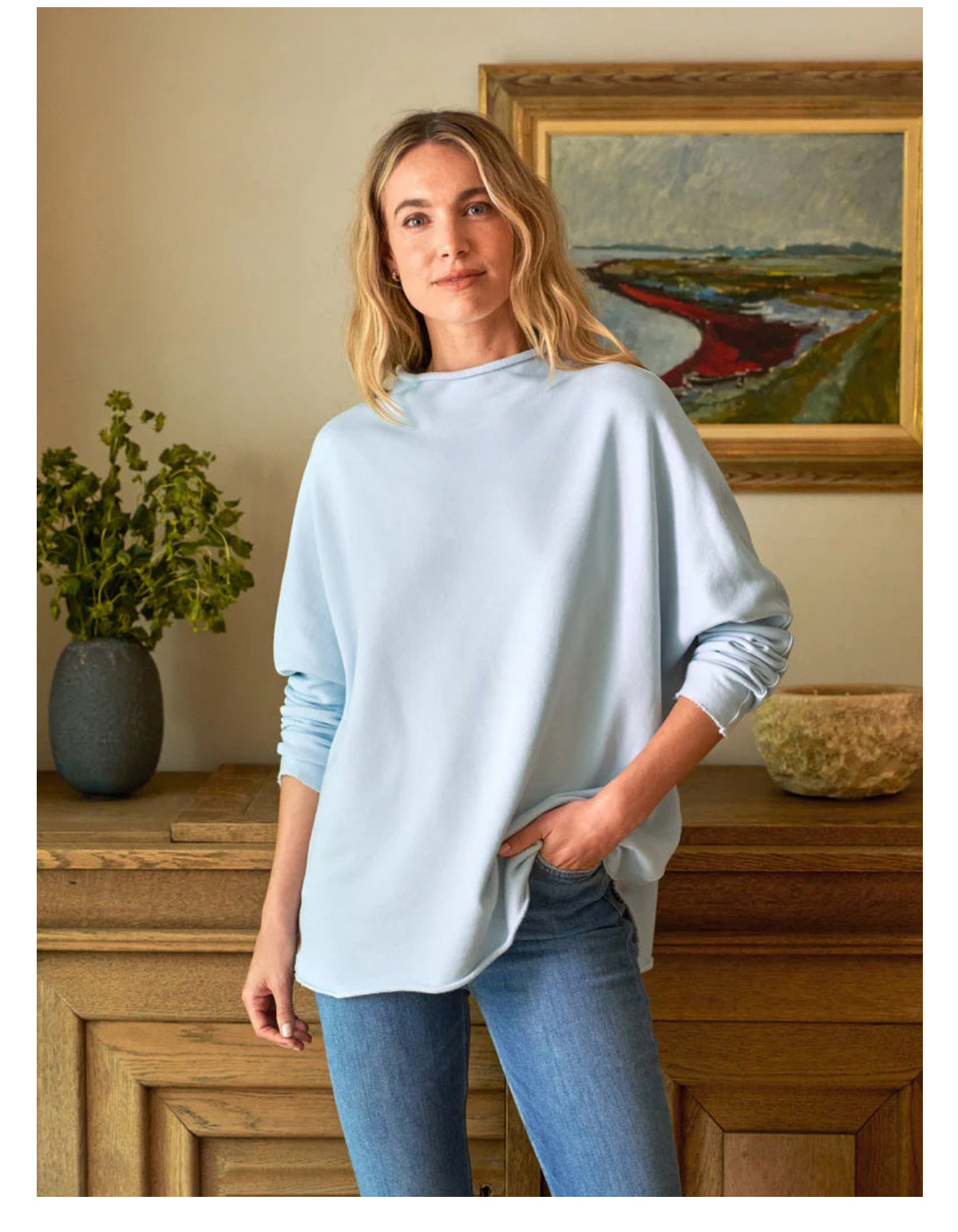 A person with long hair stands indoors wearing a light blue long-sleeve shirt and jeans. They have one hand in the pocket. A wooden table with a vase and an abstract painting on the wall is in the background. Fearrington Village