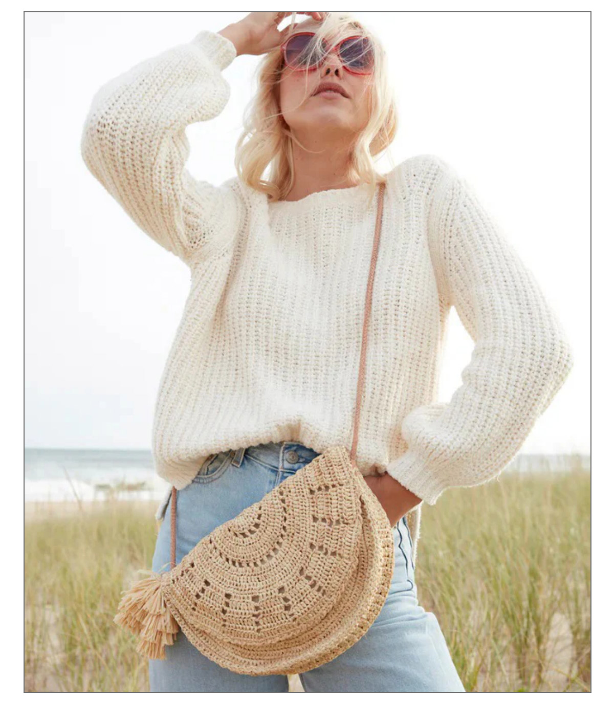 A person wearing sunglasses, a white knit sweater, and jeans stands outdoors. They have a woven crossbody bag with a tassel, and a beach scene with grass and sea is in the background. Fearrington Village