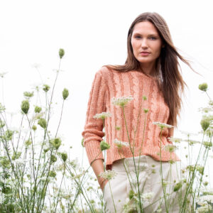 A woman stands in a field of wildflowers. She is wearing a peach-colored sweater and cream pants. The sky is overcast, creating a soft, natural backdrop. Fearrington Village