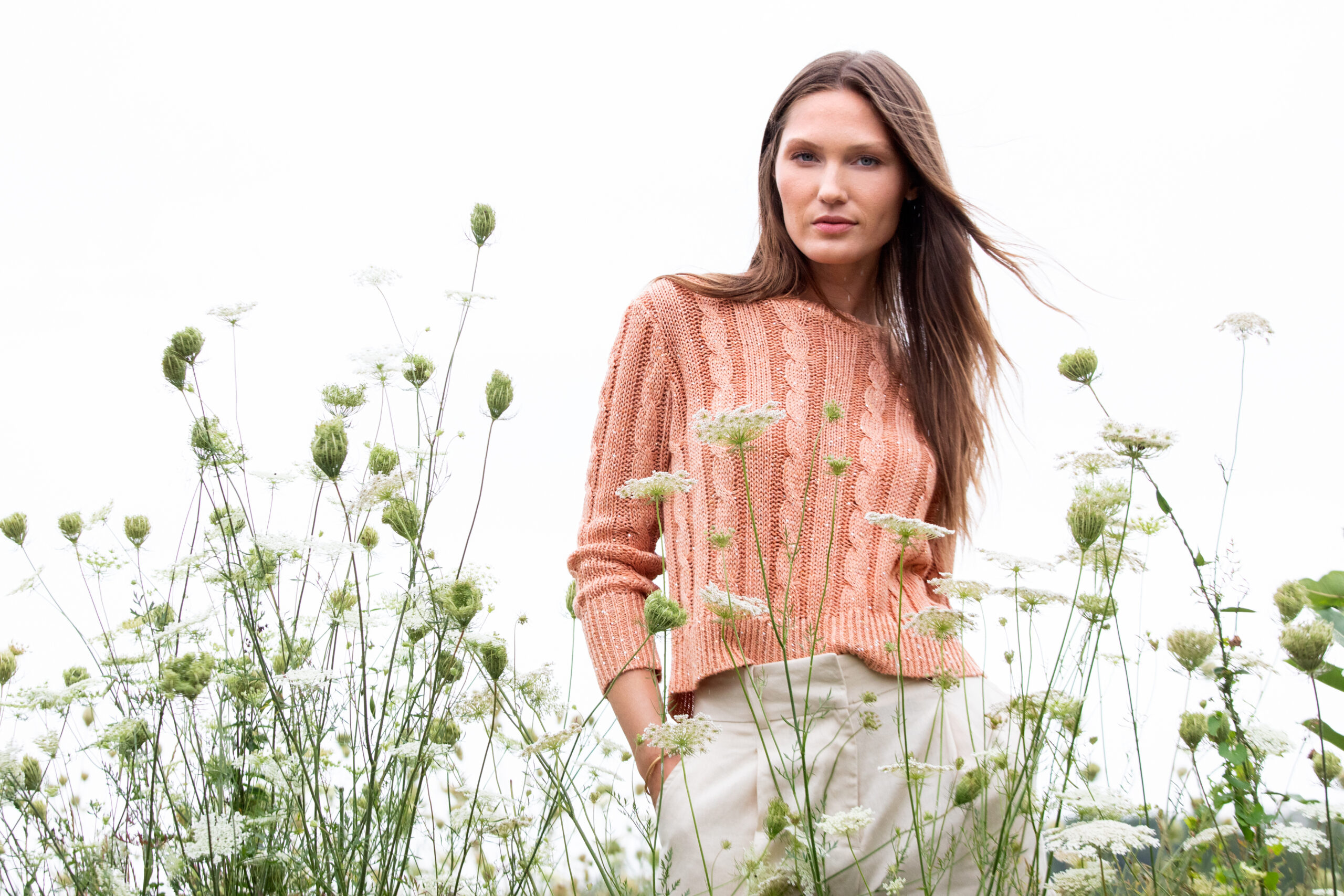 A woman stands in a field of wildflowers. She is wearing a peach-colored sweater and cream pants. The sky is overcast, creating a soft, natural backdrop. Fearrington Village