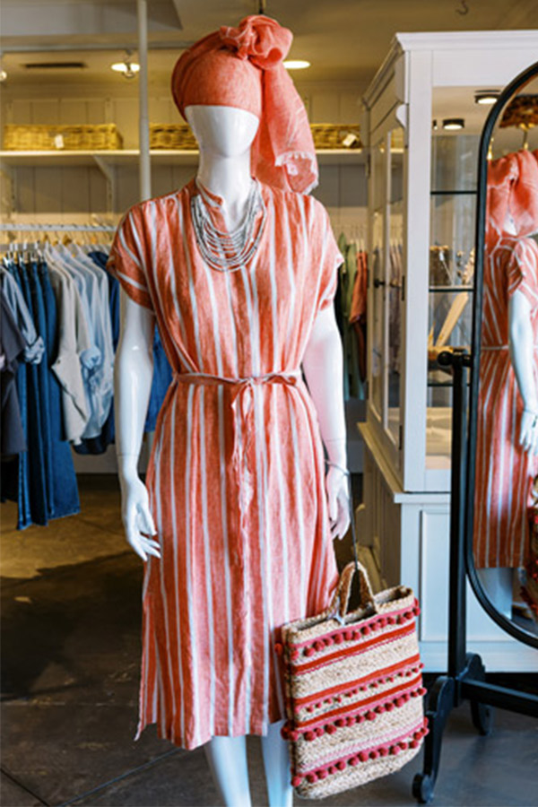 A mannequin displays a stylish outfit inside a clothing store. It wears a pink and white striped dress, a matching headwrap, and layered necklaces. A woven bag with pink accents hangs on its arm. Clothing racks and a mirror are in the background. Fearrington Village