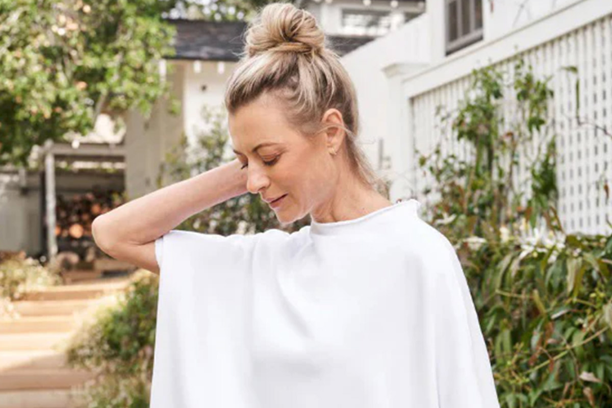 A person with light hair tied in a bun is standing outdoors, wearing a loose white top with one hand touching the back of their neck. They are in front of greenery and a white building. Fearrington Village