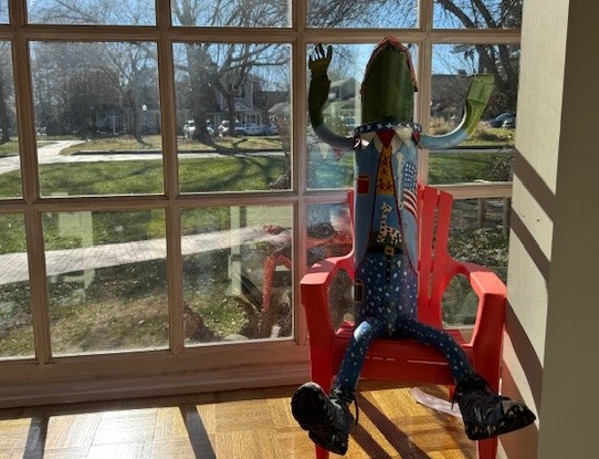 A colorful, anthropomorphic sculpture of a pickle wearing a suit and boots is sitting in a red chair by a window. The bright sun casts shadows on the wooden floor, and a grassy yard with trees is visible outside. Fearrington Village