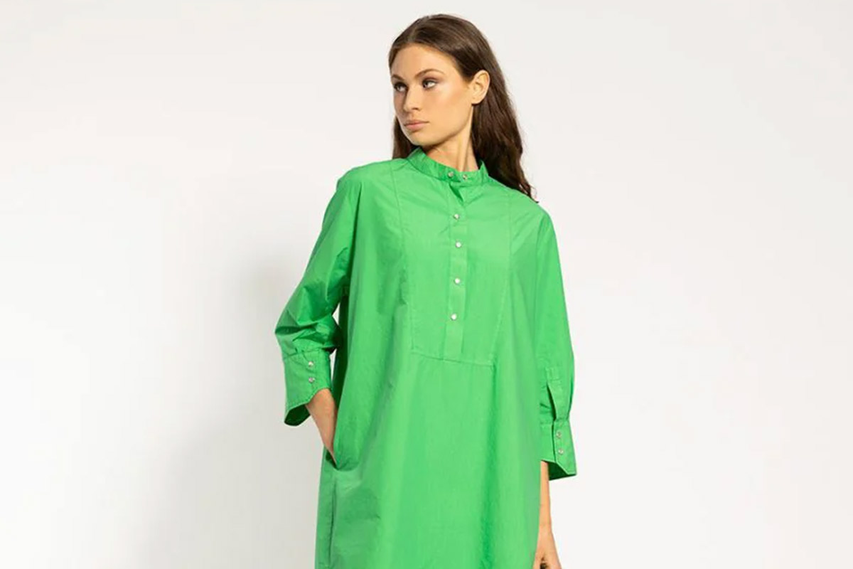 A person wearing a loose-fitting, bright green dress with long sleeves and a high collar. The dress has buttons down the front. The person is standing against a plain white background, gazing to the side with one hand in their pocket. Fearrington Village