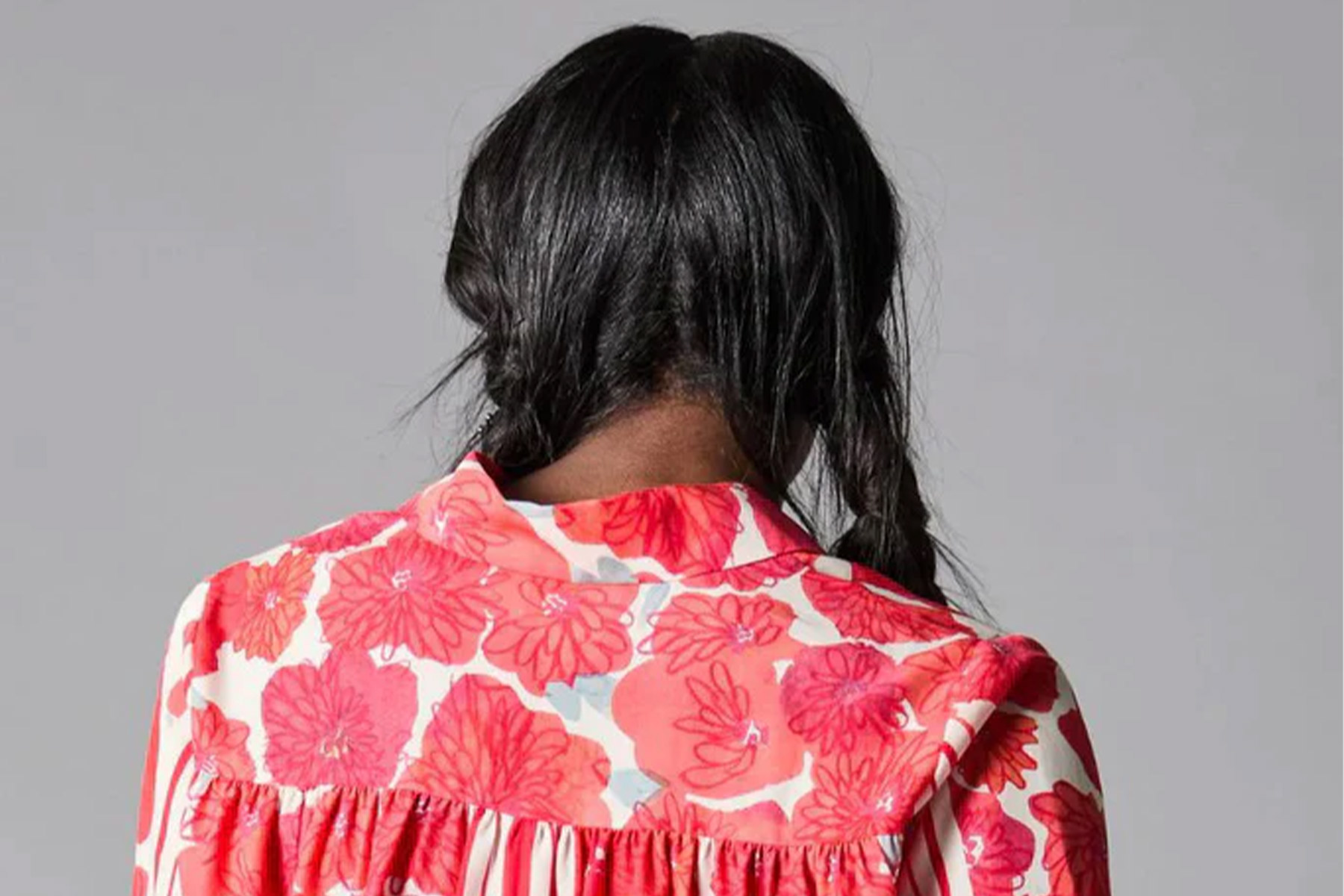 A person with long, dark hair styled in two braids is facing away from the camera. They are wearing a white blouse with a vibrant red floral pattern and a pleated back. The background is plain gray. Fearrington Village