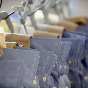 Close-up of several blue collared shirts hanging in a row, each shirt held by metal clips on hangers. The shirts are neatly arranged, displaying a uniform pattern of buttons and stitching. The background is softly blurred. Fearrington Village