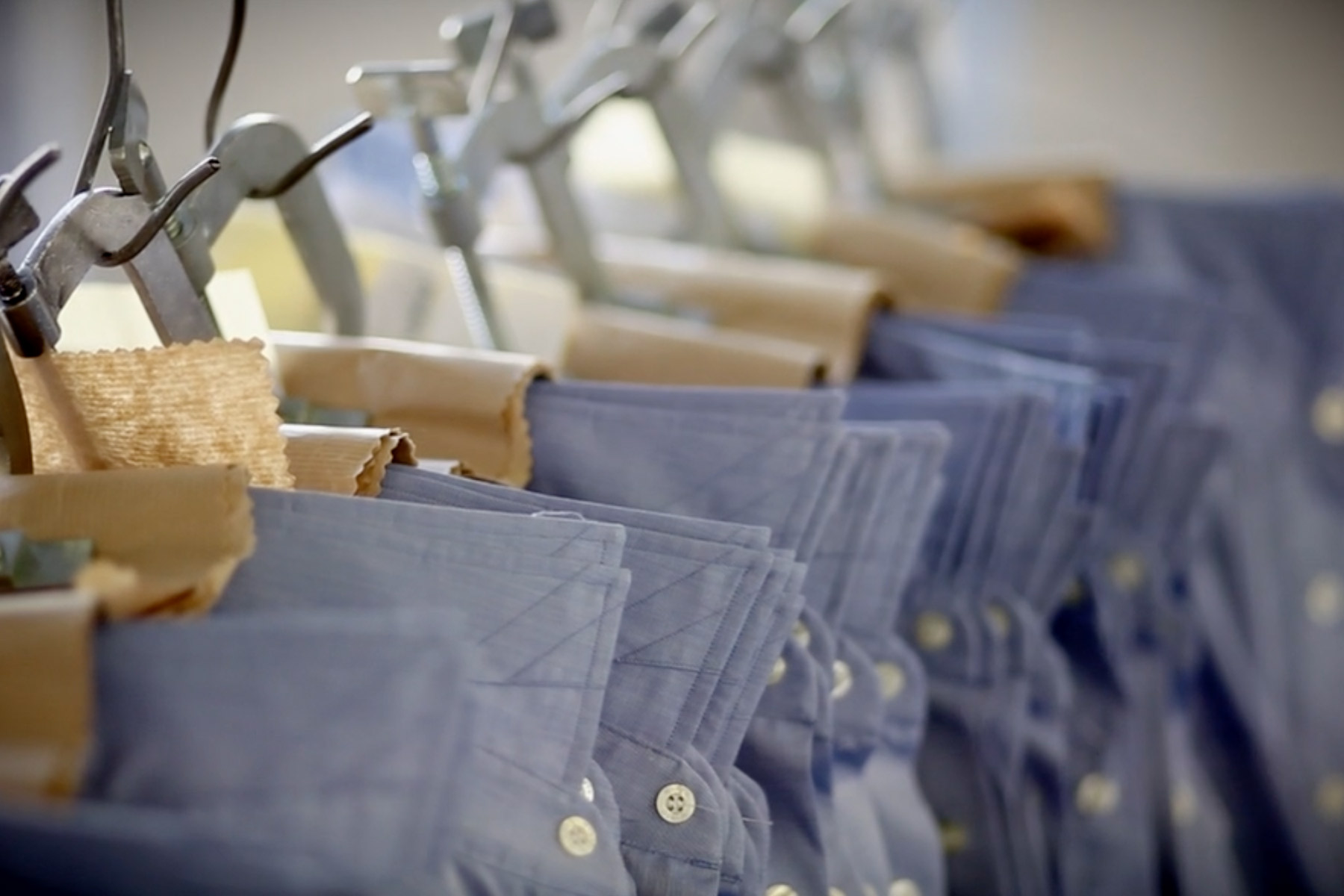 Close-up of several blue collared shirts hanging in a row, each shirt held by metal clips on hangers. The shirts are neatly arranged, displaying a uniform pattern of buttons and stitching. The background is softly blurred. Fearrington Village