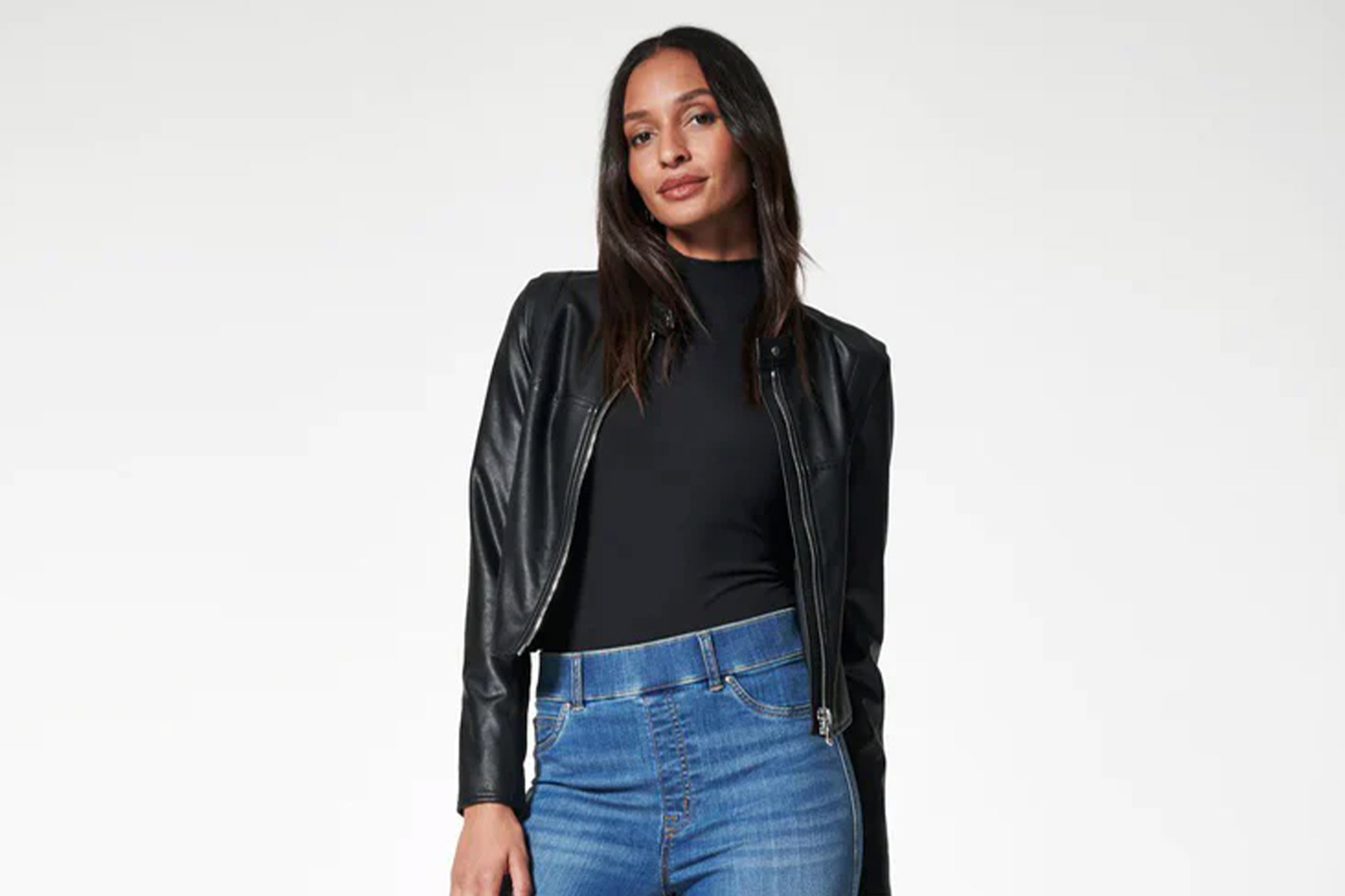 A woman with long brown hair is wearing a black leather jacket, a black top, and blue jeans. She is posing against a plain, light background. Fearrington Village