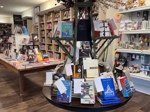 A cozy bookstore displays various books on wooden tables. The central table features staff recommendations with handwritten notes. Bookshelves and stuffed animals are in the background, creating a welcoming atmosphere. Fearrington Village