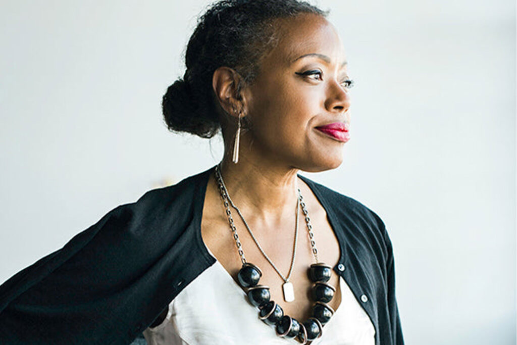 A person with short hair stands against a light background, looking into the distance. They wear a dark cardigan over a white top, a bold necklace with large beads, and have small hoop earrings. Fearrington Village