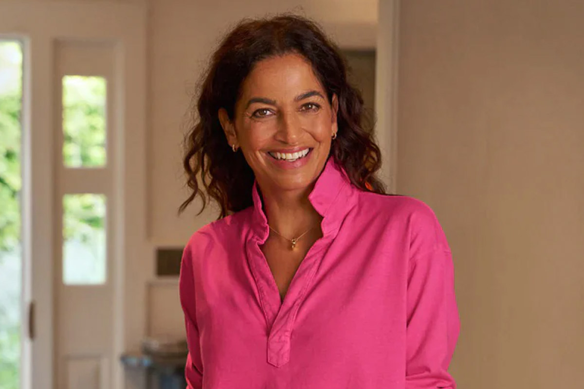 Person with wavy dark hair wearing a pink shirt, smiling and standing indoors in front of a door. Fearrington Village