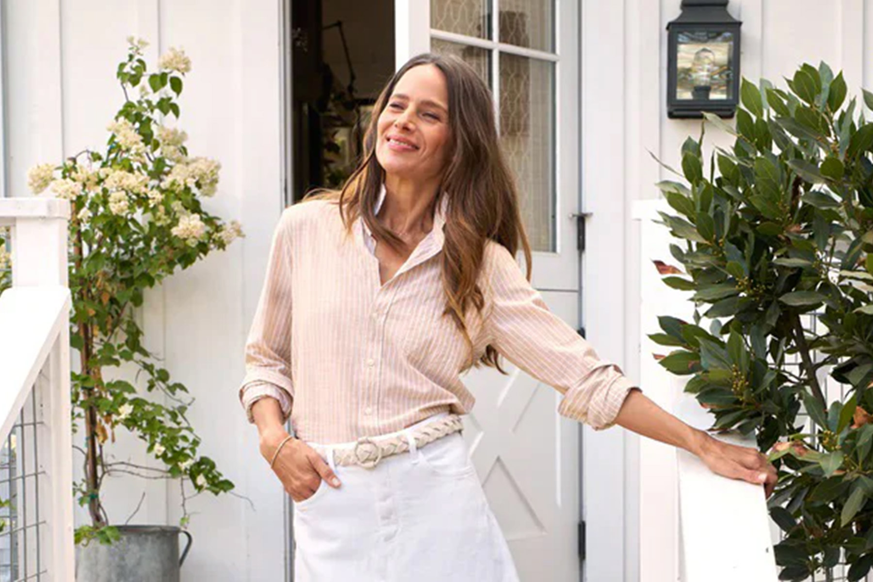 A woman is standing outside, smiling, in a garden setting. She is wearing a light-colored striped shirt and white pants. There are green plants and a white building with a door and lantern in the background. Fearrington Village