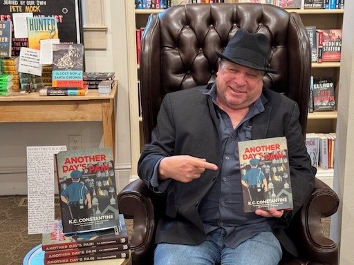 Gentle Reader - February 11, 2025 1 A man in a black hat and blue shirt sits in a leather chair in a bookstore, smiling and pointing at a book titled "Another Day's Pain" by K.C. Constantine. A stack of the books is on a table nearby. Fearrington Village