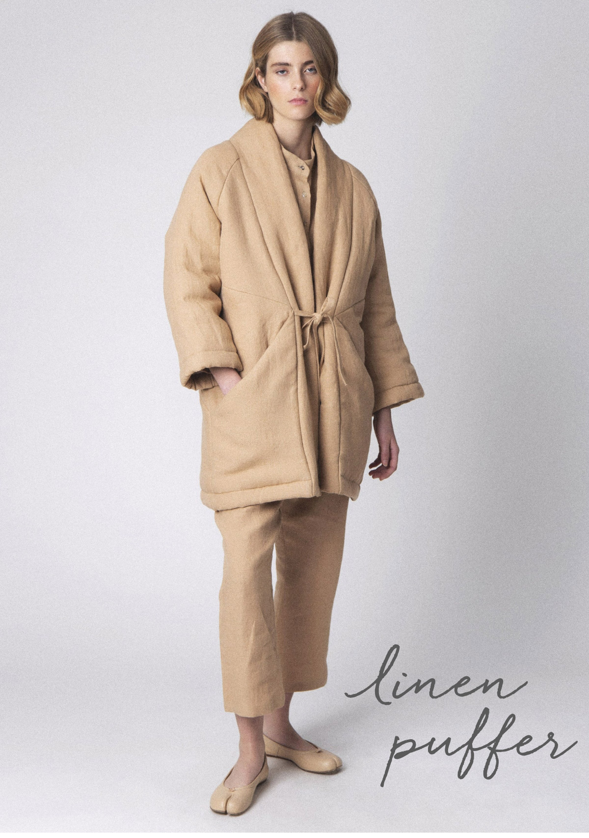 A person stands wearing a beige linen puffer coat tied at the waist, with matching pants and beige shoes. The background is plain, and the words "linen puffer" are written in cursive in the bottom right corner. Fearrington Village