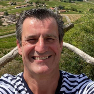 A man in a striped shirt is smiling at the camera. Behind him, there's a scenic view of a vineyard landscape with green fields, a few houses, and a clear sky. He appears to be standing on a wooden terrace or balcony. Fearrington Village