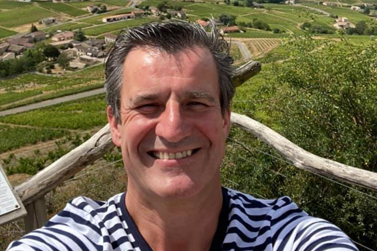 A man in a striped shirt is smiling at the camera. Behind him, there's a scenic view of a vineyard landscape with green fields, a few houses, and a clear sky. He appears to be standing on a wooden terrace or balcony. Fearrington Village