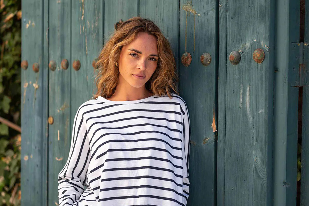 A woman with wavy hair stands against a rustic teal wooden door, wearing a white and black striped long-sleeve top. She looks at the camera with a relaxed expression. Fearrington Village