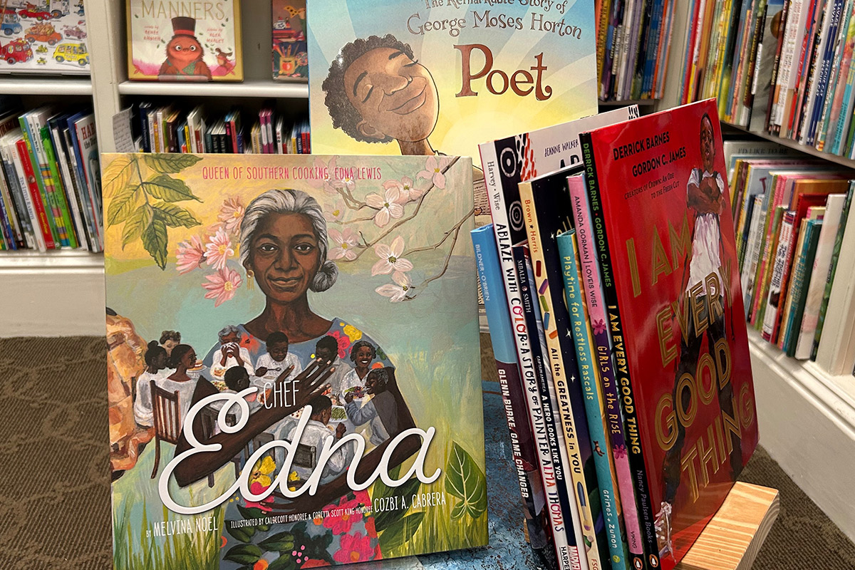 A display of children's books in a library, featuring prominently "Chef Edna" with illustrations of Edna Lewis and children, and "Poet" about George Moses Horton. The colorful covers and spines of various books fill the background. Fearrington Village