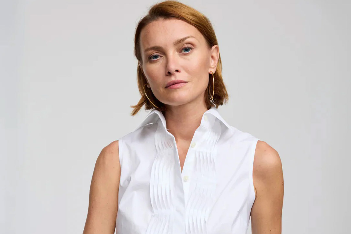 A person with short auburn hair and a neutral expression wears a sleeveless white shirt with a pleated front. They stand against a plain light background. Fearrington Village