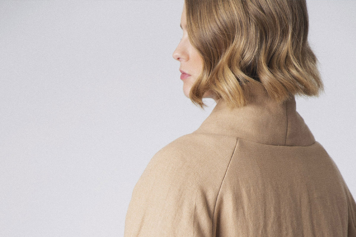 A person with wavy shoulder-length blonde hair is seen from behind, wearing a beige coat. They are standing against a plain light background. Fearrington Village