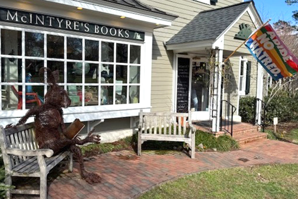 A rustic statue of a rabbit holding a book sits on a bench outside McIntyre's Books. A "No Hate" flag waves beside the bookstore entrance, which has a wooden door and brick steps. Lush greenery surrounds the building. Fearrington Village