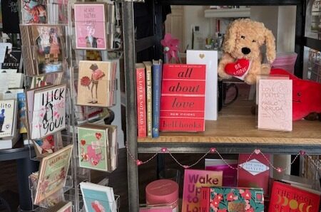 A display shelf with romantic books, greeting cards, and a plush dog holding a heart that reads "Be Mine." The main book, "All About Love" by bell hooks, is centered. Decorative heart-themed accents surround the items. Fearrington Village