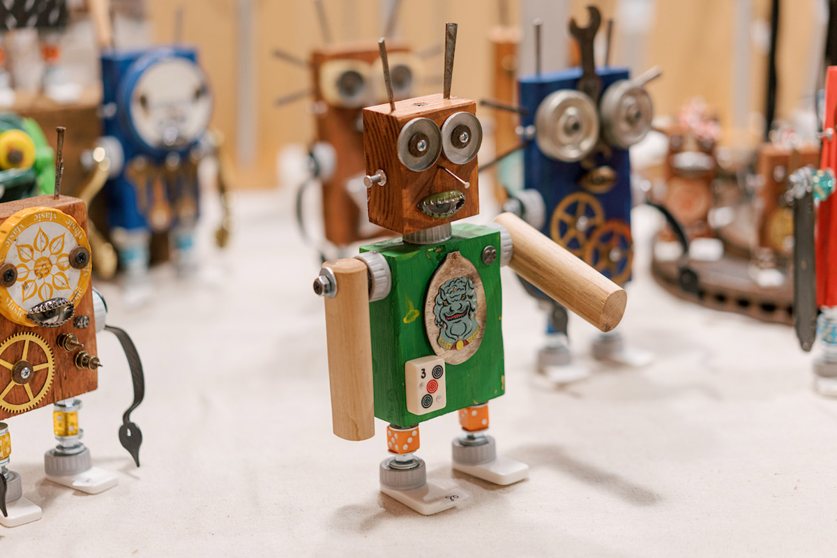 A small, colorful wooden robot with a green body, round eyes, and a quirky expression stands on a table. It has various gears and mechanical parts. Other similar robots are blurred in the background. Fearrington Village