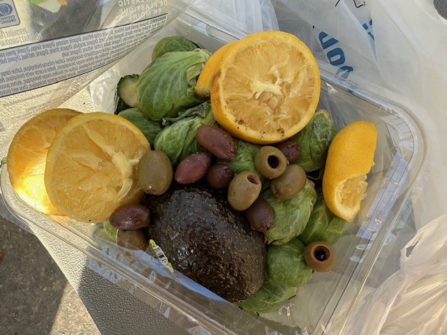 A clear plastic container filled with Brussels sprouts, a halved avocado, and a mix of green and black olives. Slices of lemon and a halved orange are also placed on top. The container is set on a gray surface. Fearrington Village