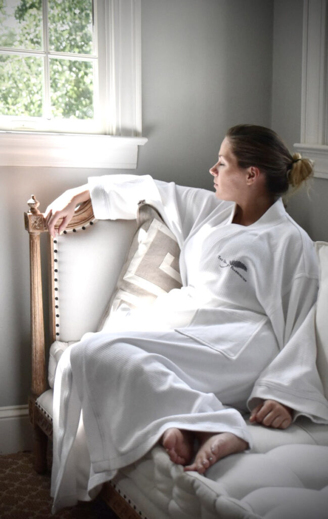 A person in a white robe sits on a cushioned chair by a window, gazing outside. Sunlight illuminates their relaxed posture. The room has neutral tones, and a decorative pillow adds a touch of pattern to the scene. Fearrington Village