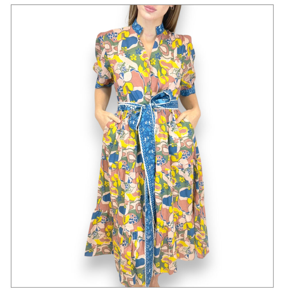 A woman wearing a colorful dress with a vibrant abstract pattern stands with her hands in the dress pockets. The dress features a blue belt and a high collar. The background is plain white. Fearrington Village