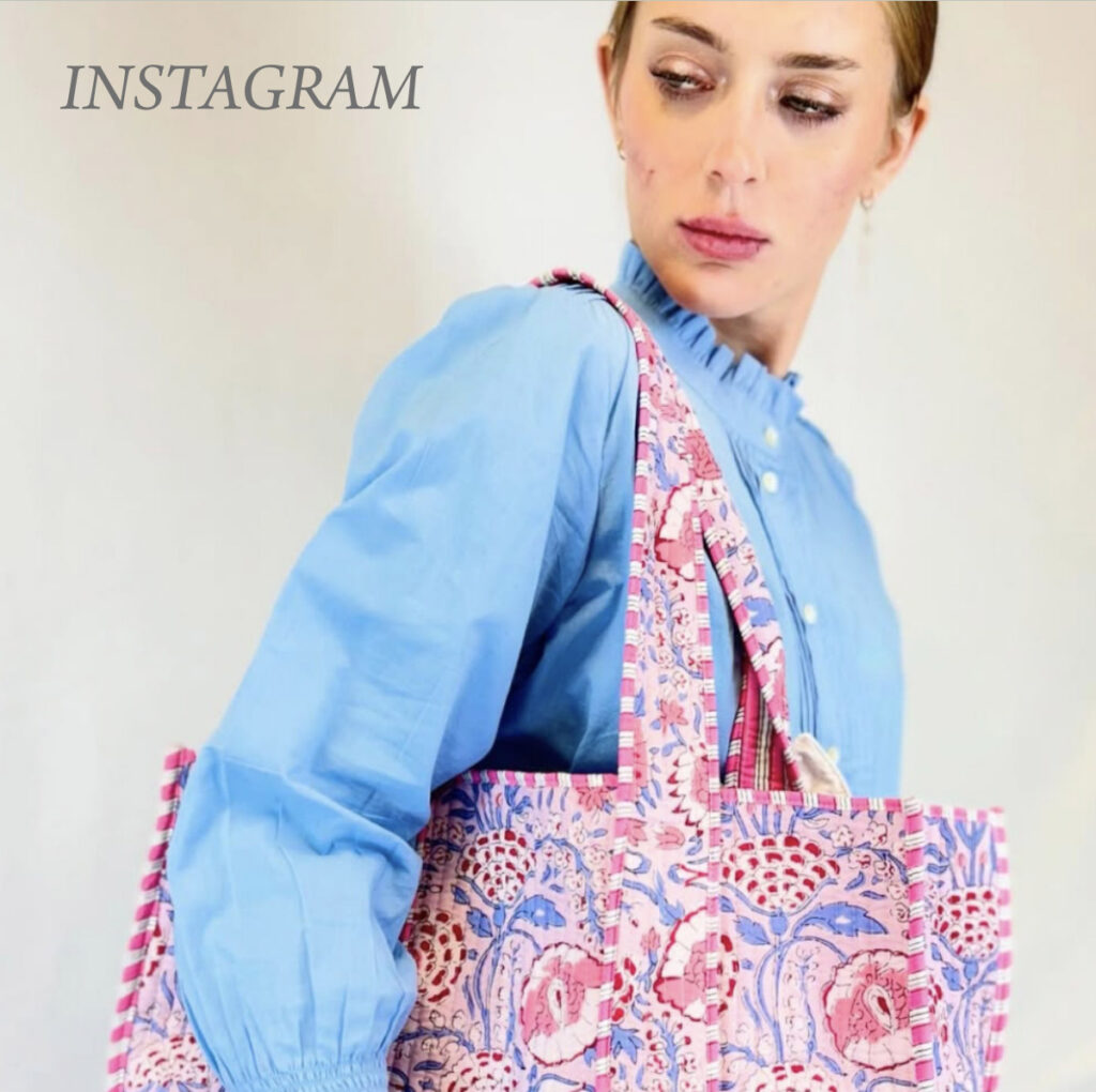 A person wearing a light blue blouse with ruffled collar looks to their side while carrying a colorful floral-patterned tote bag on their shoulder. The word "Instagram" is written in the top left corner. Fearrington Village