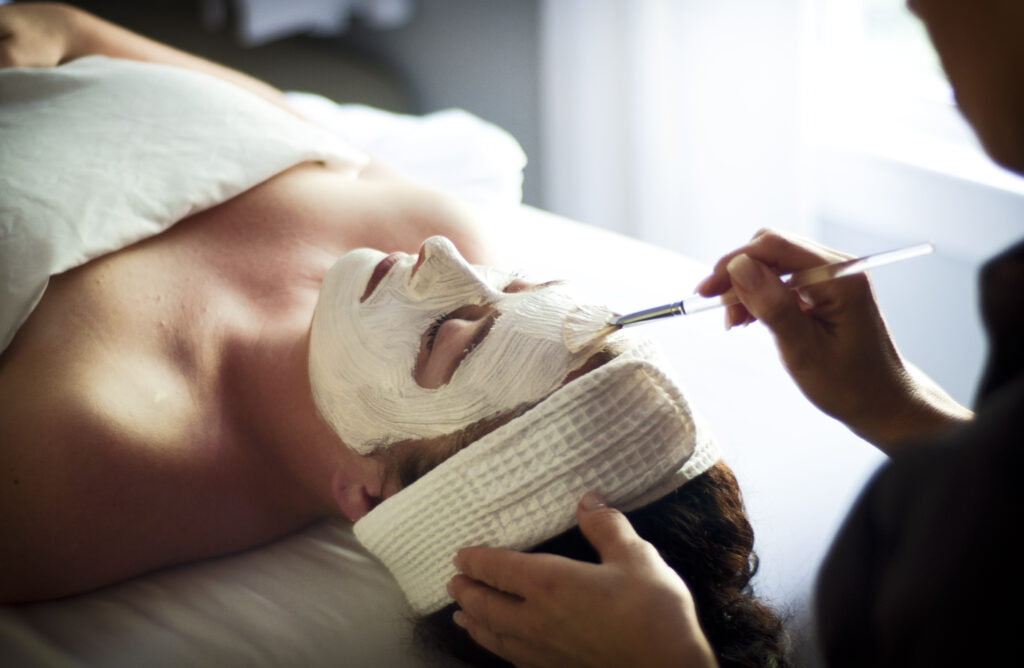 Person lying on a spa table with a towel on their head, receiving a facial treatment. Another person applies a white face mask with a brush. The setting is calm and relaxing, with soft lighting. Fearrington Village