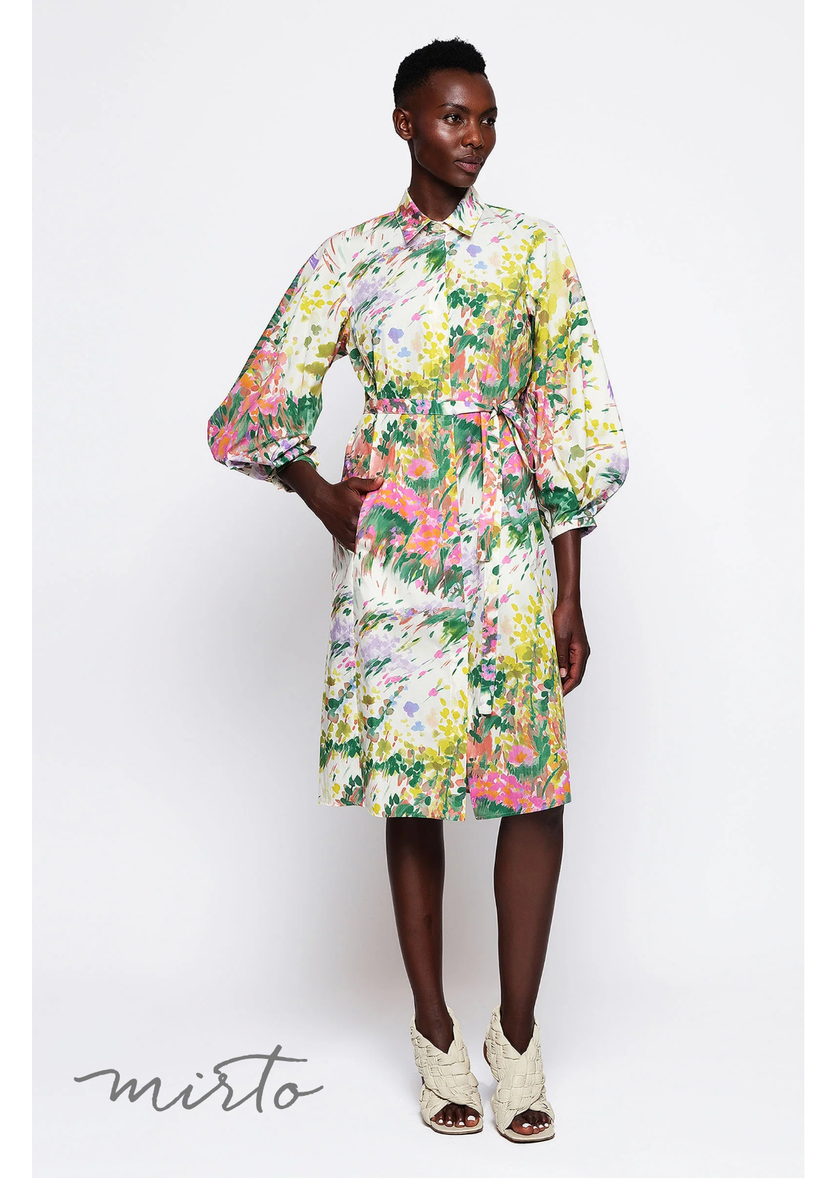 A person is wearing a colorful, floral-patterned dress with three-quarter sleeves and belted waist. They stand in front of a white background, wearing cream-colored peep-toe shoes. The word "mirto" is at the bottom left corner. Fearrington Village
