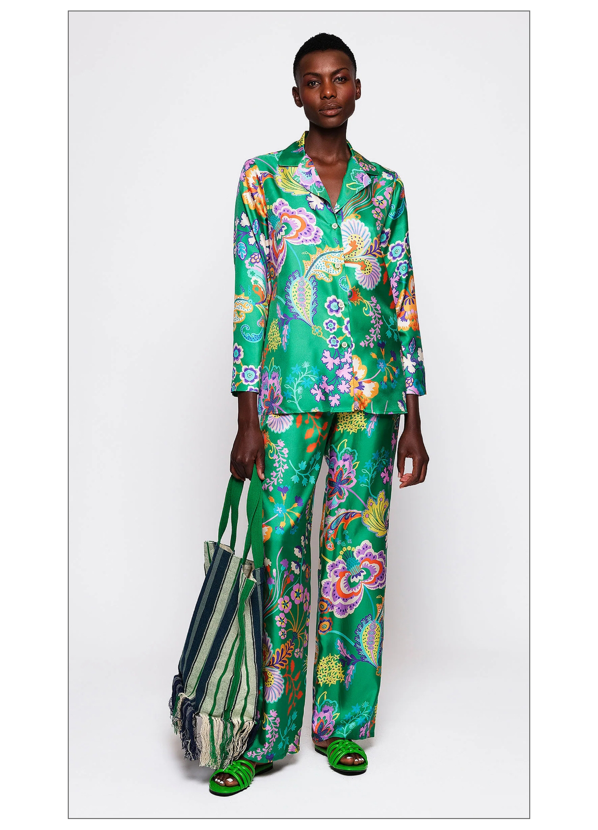 A person poses in a vibrant green satin outfit with colorful floral patterns. They are holding a large striped tote bag with fringes and wearing matching sandals. The background is plain white. Fearrington Village