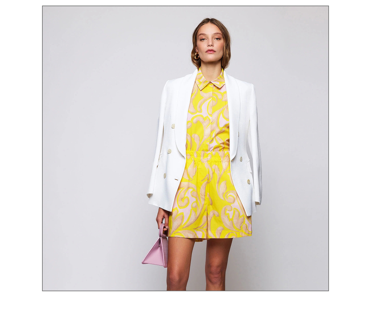 A woman stands against a neutral background wearing a white blazer over a yellow patterned shirt and shorts. She carries a small purple handbag and has straight hair. Fearrington Village