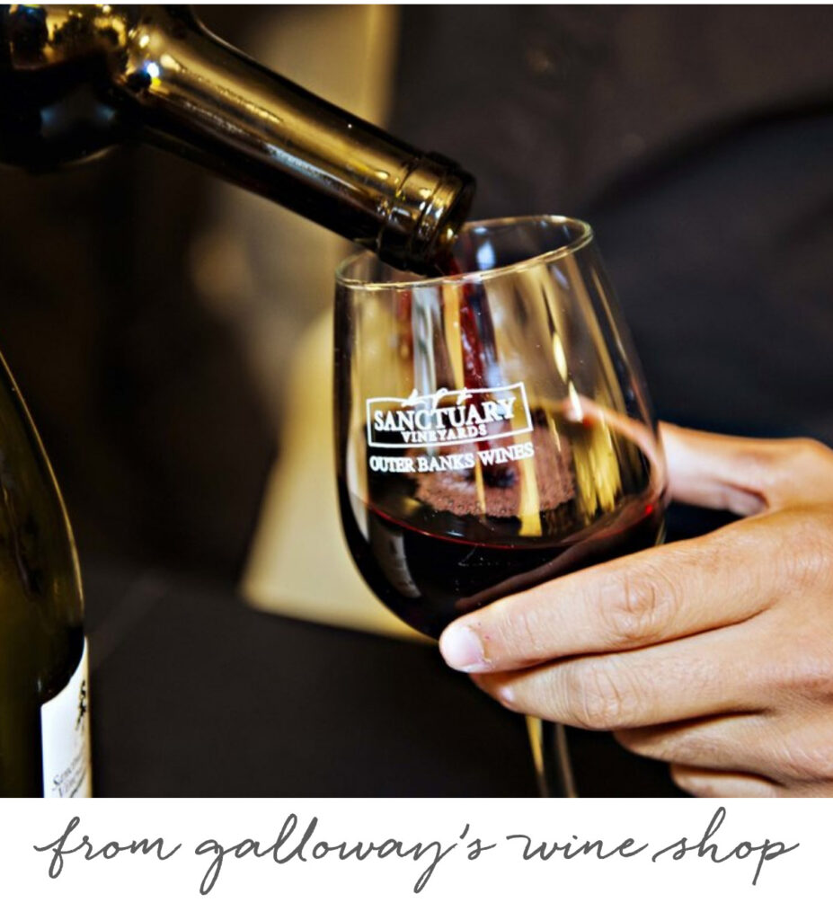 A person pours red wine into a branded wine glass. The glass reads "Sanctuary Vineyards Outer Banks Wines." The background is blurred, and text below reads "from galloway's wine shop. Fearrington Village
